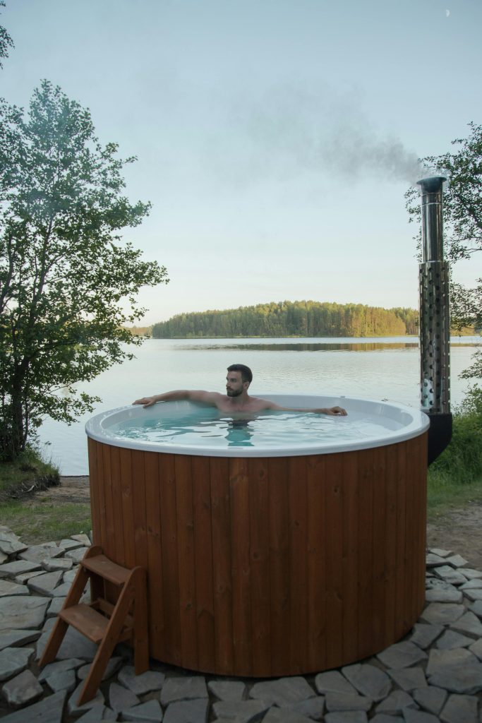 Shirtless man enjoying relaxation in a hot tub outdoors by a serene lake.